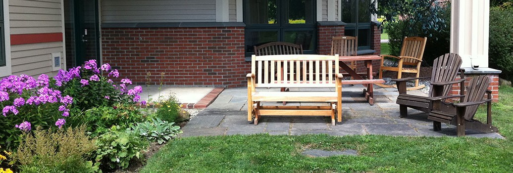 a bench lit up by sunshine. The library building is visible behind it and there are several other chairs around it as well as flowers growing nearby /end ID