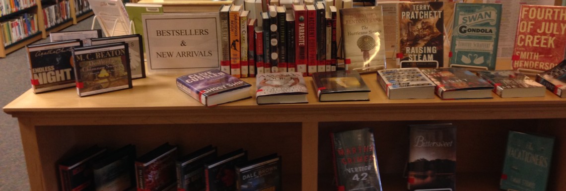a display of new books on a waist height bookshelf. There are books on the shelf and on top. There is a sign that says "Bestsellers and New Arrivals" /end ID