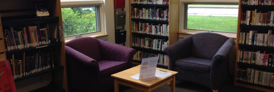 the YA nook. There are two purple chairs and a small table, with 3 bookshelves filled with books lining the walls /end ID